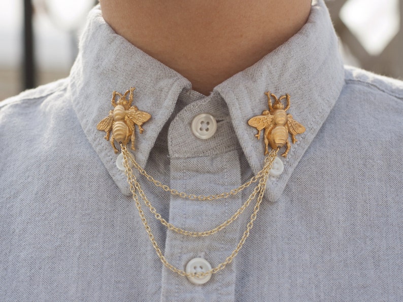 May include: Gold-colored bee collar clips with dangling chains, attached to a light gray button-down shirt. The bee-shaped clips are detailed with wings and body segments. The chains hang below the shirt collar.