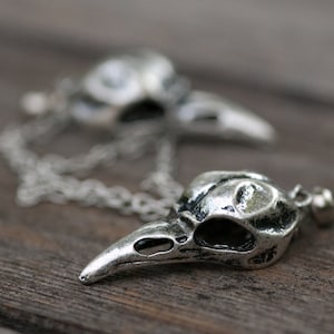 May include: A silver chain necklace with two pendant charms shaped like bird skulls. The charms are detailed and have a textured finish.