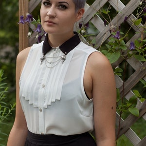 May include: A woman with short, lavender-colored hair is wearing a white blouse with a black Peter Pan collar and a silver chain necklace with a decorative pendant. She is also wearing a black skirt.