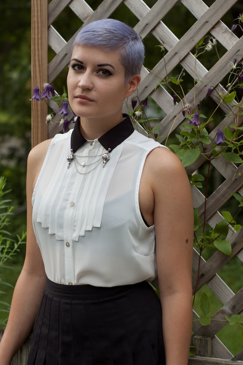 May include: A woman with short, lavender-colored hair is wearing a white blouse with a black Peter Pan collar and a silver chain necklace with a decorative pendant. She is also wearing a black skirt.
