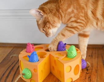 May include: A playful orange tabby cat interacts with a yellow cheese-shaped cat toy. The toy features colorful mouse-shaped pieces in pink, blue, green, and purple. The cat's paw is reaching for one of the toy mice.