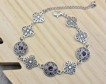 May include: A silver-toned metal bracelet featuring alternating circular and quatrefoil-shaped links. The circular links contain a dark red center surrounded by small gray petals, while the quatrefoil links feature an intricate openwork design.