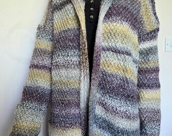 May include: A long-sleeved hooded cardigan featuring a variegated knit pattern in shades of purple, yellow, gray, and white. The garment has an open front design and is displayed on a mannequin wearing a black shirt with a button-up neckline.