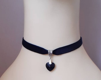 May include: A black velvet choker with a silver heart pendant featuring a black crystal. The choker is on a white mannequin.