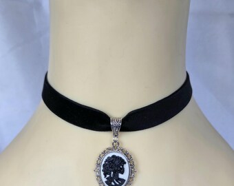 May include: Black velvet choker necklace with a silver-toned pendant. The pendant features a black and white cameo design within an oval frame. The cameo depicts a woman's profile.
