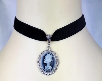 May include: Black velvet choker necklace with a silver pendant. The pendant features a cameo of a skeleton wearing a top hat. The oval pendant is framed with a decorative silver border. The necklace is displayed on a mannequin.