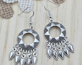 May include: A pair of silver-toned earrings. Each earring features a circular design with star-shaped cutouts and dangling teardrop-shaped charms. The earrings have a hook closure and are displayed on a wooden surface.