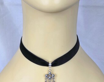 May include: Black velvet choker necklace with a silver pendant featuring skulls and three silver spikes. The necklace is worn around the neck.