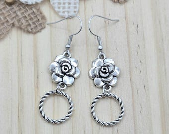 May include: Silver-toned dangle earrings. Each earring features a detailed flower design at the top, connected to a twisted rope-like circle. The earrings hang from a simple hook. The background is a light wood surface.