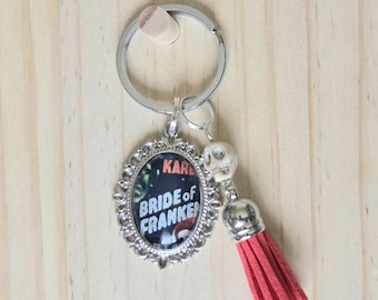 May include: A silver-toned keychain featuring an oval pendant with a Bride of Frankenstein graphic, a small white skull bead, and a red suede tassel. The pendant has an ornate, scalloped metal frame and is attached to a standard circular metal ring.