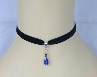 May include: A black velvet choker necklace with a silver pendant and a teardrop-shaped blue crystal. The necklace is displayed on a mannequin, showcasing the elegant design and color contrast.