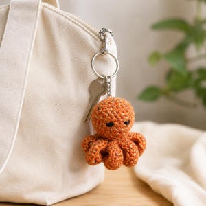 May include: A small, crocheted octopus keychain in a speckled orange yarn with dark embroidered eyes. The octopus is attached to a silver-toned metal split ring and a lobster clasp, which is clipped onto the zipper pull of a cream-colored canvas bag.