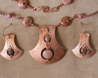 May include: A copper necklace and earrings set with pink and brown stones. The necklace features a large pendant with a hammered copper design and a pink stone in the center. The earrings are smaller and have a similar design with a brown stone in the center.