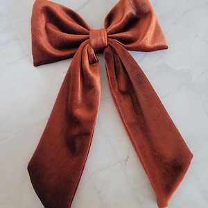 May include: A large, burnt orange velvet bow with long, draping tails. The bow is knotted in the centre and has a soft, plush texture. This hair accessory is ideal for adding a touch of elegance to any hairstyle.