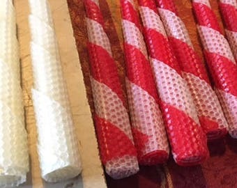 May include: A collection of red and white striped beeswax candles and plain white beeswax candles. The candles are arranged on a table with a red and white checkered tablecloth.