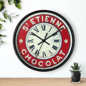 Puede incluir: Un reloj con marco negro y una esfera roja y blanca. La esfera del reloj tiene números romanos y las palabras "St. Etienne Chocolat" alrededor del borde.
