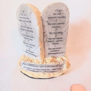 May include: A decorative religious figurine depicting two stone tablets with the Ten Commandments inscribed in black text. The base includes a quote from John 13:34. The tablets are beige with a textured appearance.