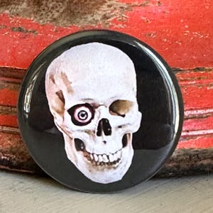Könnte beinhalten: Ein runder Ansteckbutton mit der Illustration eines weißen menschlichen Schädels vor einem einfarbigen schwarzen Hintergrund. Der Schädel zeigt ein einzelnes sichtbares Auge mit detaillierter Iris und Pupille, während die andere Augenhöhle leer bleibt.