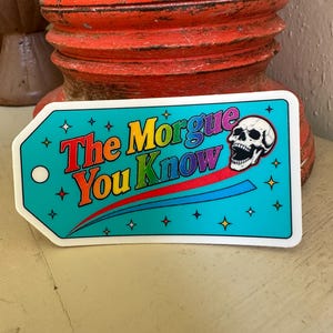 Puede incluir: Una pegatina rectangular con forma de etiqueta con un fondo azul turquesa brillante que presenta el texto The Morgue You Know en una fuente degradada multicolor. Una calavera blanca con la boca abierta se sitúa a la derecha, rodeada de pequeñas estrellas amarillas.