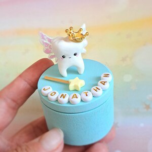 May include: A light blue cylindrical wooden box featuring a white tooth figurine with iridescent wings and a gold crown on the lid. A yellow star wand and white beads spelling the name JONATHAN are attached to the top of the lid.