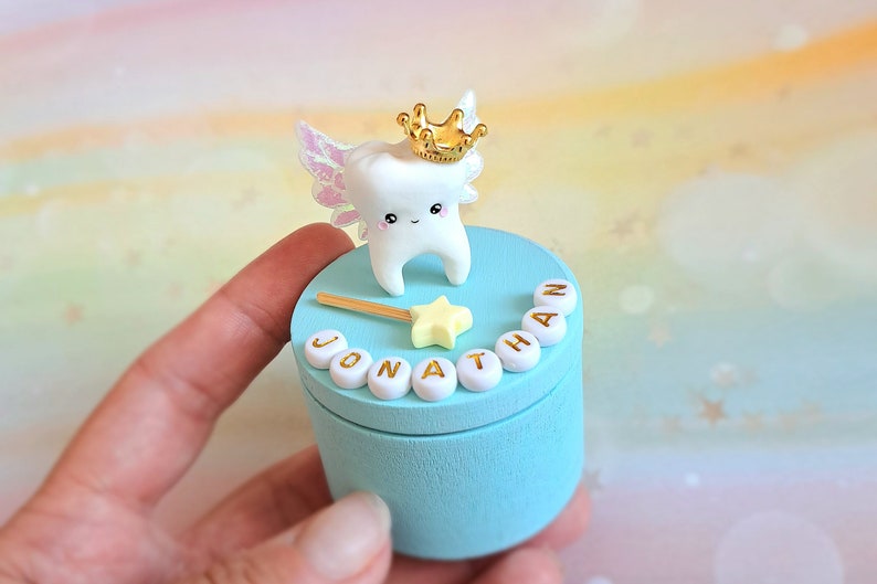 May include: A light blue cylindrical wooden box featuring a white tooth figurine with iridescent wings and a gold crown on the lid. A yellow star wand and white beads spelling the name JONATHAN are attached to the top of the lid.
