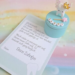 May include: A light blue round keepsake box topped with a white tooth figurine wearing a gold crown and iridescent wings, featuring a pink star wand and the name ELENI in white beads. Beside it lies a card with German text and a tooth graphic.