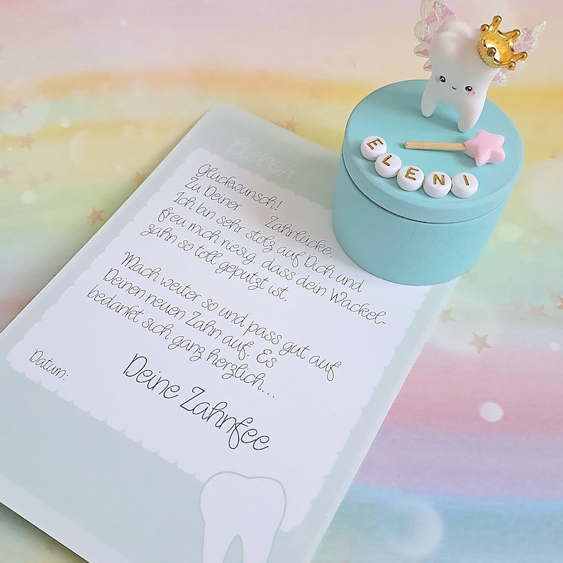 May include: A light blue round keepsake box topped with a white tooth figurine wearing a gold crown and iridescent wings, featuring a pink star wand and the name ELENI in white beads. Beside it lies a card with German text and a tooth graphic.