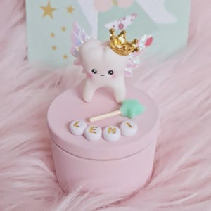 May include: A round light pink storage box featuring a white tooth figurine with iridescent wings and a gold crown on the lid. A small mint green star wand and four white beads spelling the name LENI in gold letters are attached to the lid.