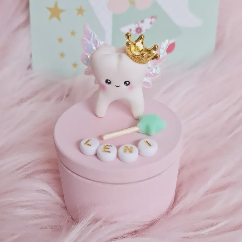 May include: A round light pink storage box featuring a white tooth figurine with iridescent wings and a gold crown on the lid. A small mint green star wand and four white beads spelling the name LENI in gold letters are attached to the lid.