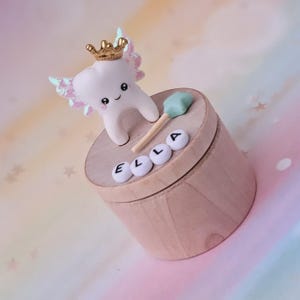 May include: A round wooden box featuring a white tooth figurine with iridescent wings and a gold crown on its head. A light green star-shaped wand rests on the lid next to white letter beads spelling the name ELLA in black capital letters.