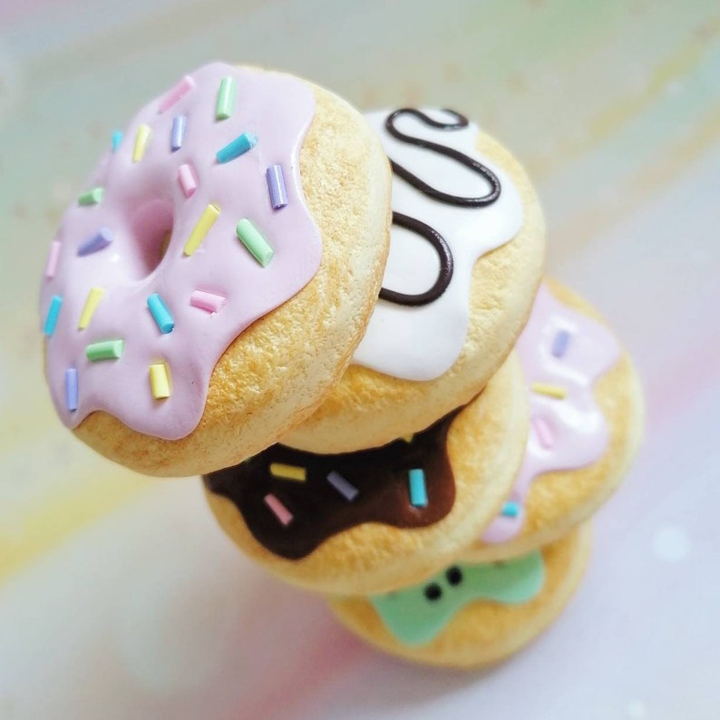 May include: A stack of four mini donuts with different colored icings and sprinkles. The top donut is pink with colorful sprinkles, the second is white with black swirls, the third is brown with pink and blue sprinkles, and the bottom is green with black dots.