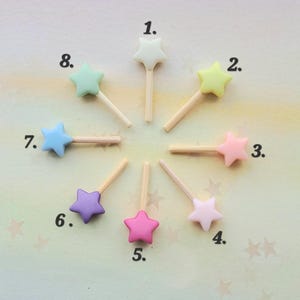 May include: Eight star-shaped toppers on wooden sticks arranged in a circle, numbered 1 through 8. The stars are in various pastel colors including white, yellow, pink, light pink, light purple, purple, blue, and mint green, each with a wooden stem.