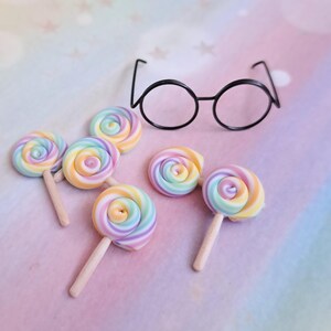 May include: Six rainbow swirl lollipops on sticks. The lollipops are arranged in a semi-circle on a pink and blue background.