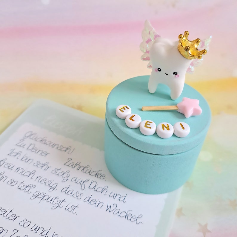 May include: A round light blue wooden box featuring a white tooth figurine with iridescent wings and a gold crown on the lid. The name ELENI is spelled out in white letter beads, accompanied by a small pink star wand and a card with German text.