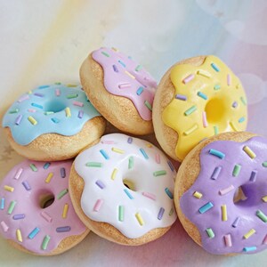 May include: Six colorful mini donuts with pastel frosting and sprinkles. The donuts are arranged in a circle on a light pink background.