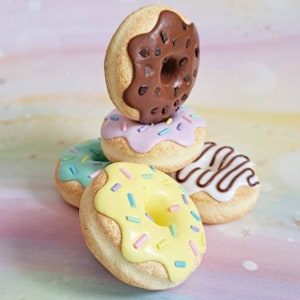 May include: A stack of four colorful mini donuts with different toppings, including chocolate, sprinkles, and icing. The donuts are made of polymer clay and are perfect for a miniature food collection.