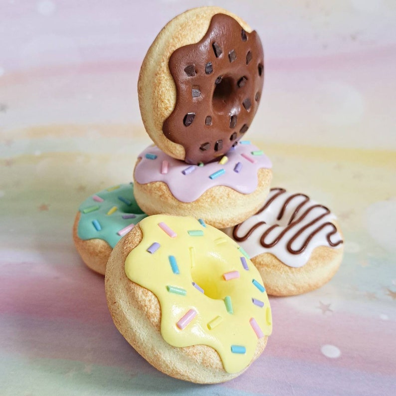 May include: A stack of four colorful mini donuts with different toppings, including chocolate, sprinkles, and icing. The donuts are made of polymer clay and are perfect for a miniature food collection.