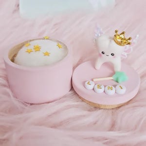 May include: A round pink container with a white cushion top decorated with small gold stars. Beside it, a pink lid features a white tooth figurine wearing a gold crown with iridescent wings, a small green star wand, and white beads spelling LENI.