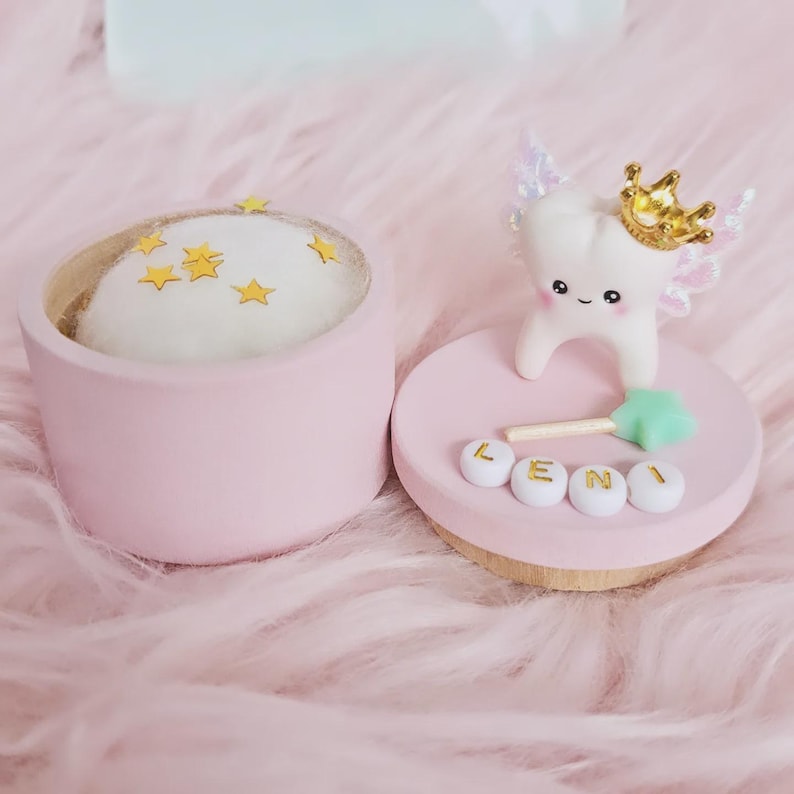 May include: A round pink container with a white cushion top decorated with small gold stars. Beside it, a pink lid features a white tooth figurine wearing a gold crown with iridescent wings, a small green star wand, and white beads spelling LENI.