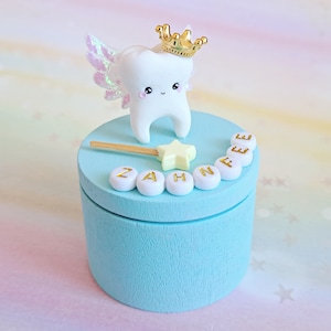May include: A light blue cylindrical wooden box featuring a white tooth figurine with iridescent wings and a gold crown on its lid. A small yellow star wand and white beads with the gold text ZAHNFEE are arranged on the top surface of the box.