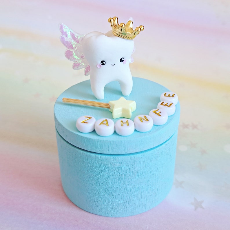 May include: A light blue cylindrical wooden box featuring a white tooth figurine with iridescent wings and a gold crown on its lid. A small yellow star wand and white beads with the gold text ZAHNFEE are arranged on the top surface of the box.