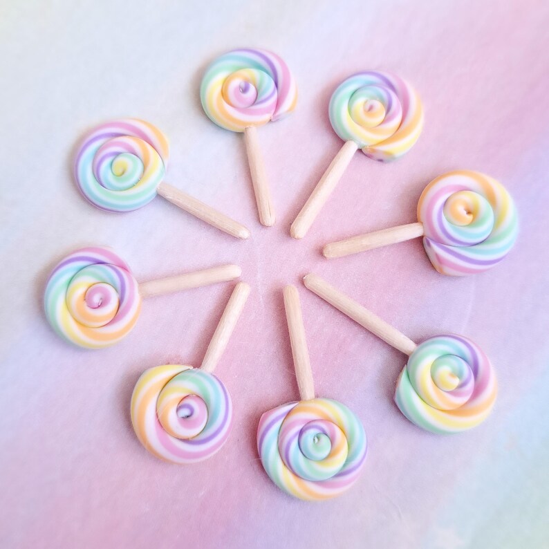 May include: Eight rainbow swirl lollipops on wooden sticks arranged in a circle on a pink and purple ombre background.
