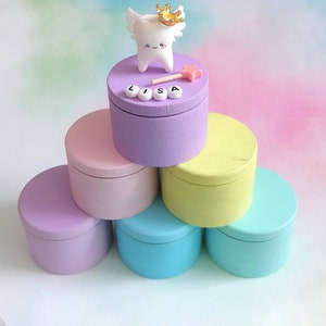 May include: A stack of six round wooden boxes in pastel colors including purple, pink, yellow, and blue. The top purple box features a white tooth figurine with wings and a gold crown, a small pink star wand, and white letter beads spelling LISA.