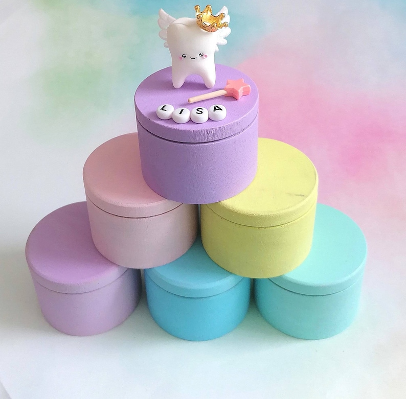 May include: A stack of six round wooden boxes in pastel colors including purple, pink, yellow, and blue. The top purple box features a white tooth figurine with wings and a gold crown, a small pink star wand, and white letter beads spelling LISA.