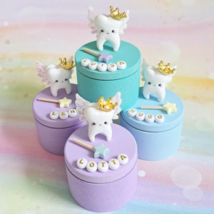 May include: Four round wooden keepsake boxes in teal, light blue, and lavender, each topped with a white tooth figurine wearing a gold crown and iridescent wings. Each box features a small star wand and white letter beads spelling LUISA, LEN, HUGO, or LOTTA.