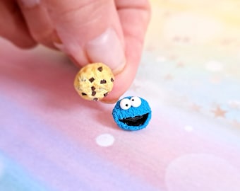 Könnte beinhalten: Ein Miniatur-Schokoladen-Chip-Keks und eine blaue, pelzige Figur mit offenem Mund und Kulleraugen. Der Keks wird von den Fingern einer Person gehalten, und die Figur liegt auf einer bunten Oberfläche.