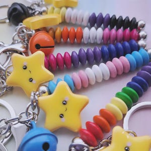 Könnte beinhalten: Eine Sammlung bunter Schlüsselanhänger mit Perlen und Stern- und Glockenanhängern. Die Schlüsselanhänger weisen eine Vielzahl leuchtender Farben auf, darunter Gelb, Orange, Rosa, Blau und Grün. Die Perlen sind mit silberfarbenen Metallketten aufgereiht.