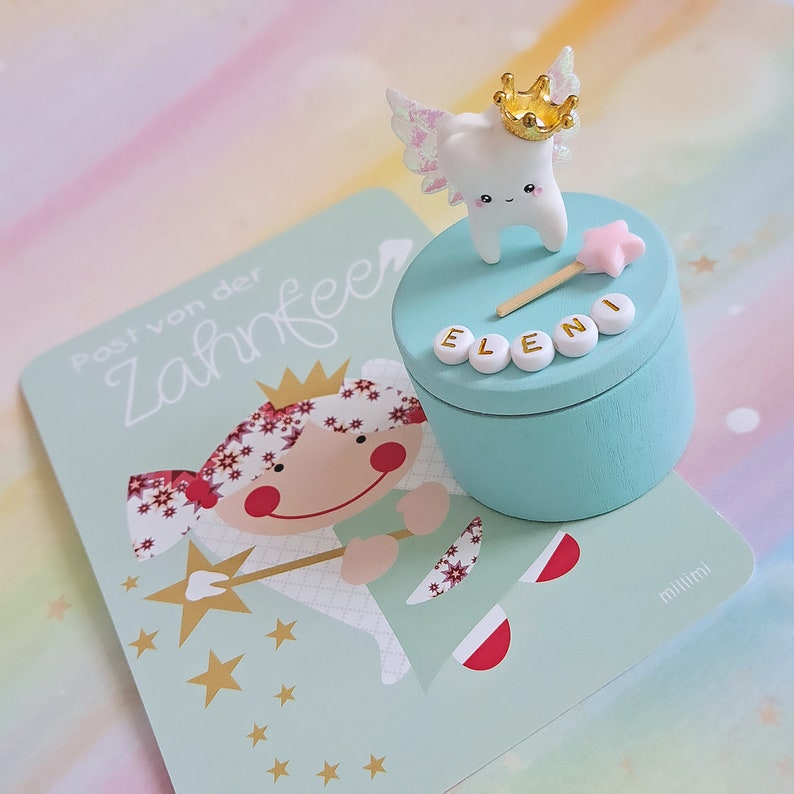 May include: A light blue round wooden box with a white tooth figurine wearing a gold crown and iridescent wings on the lid. A pink star wand and white letter beads spelling ELENI are on the lid. Beside it is a card with the text Post von der Zahnfee.