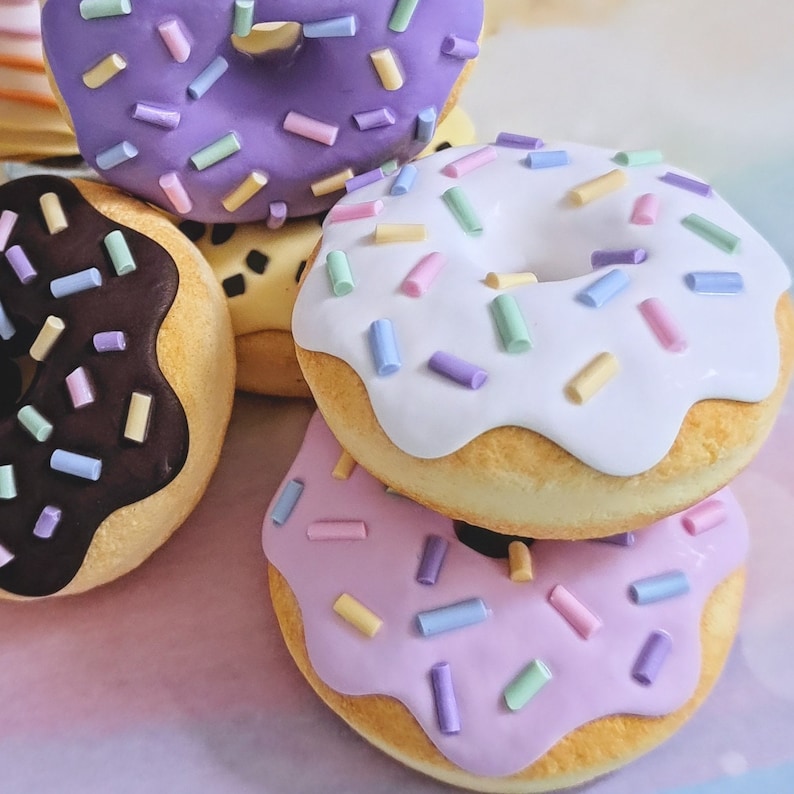 May include: A stack of four mini donuts with different colored frosting and sprinkles. The donuts are purple, white, brown, and pink.