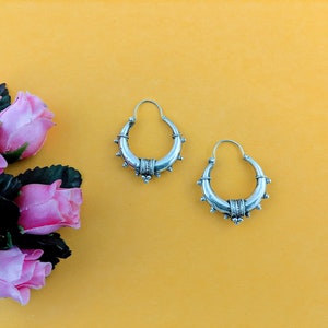 May include: A pair of silver hoop earrings with a decorative design. The earrings are on a yellow background.